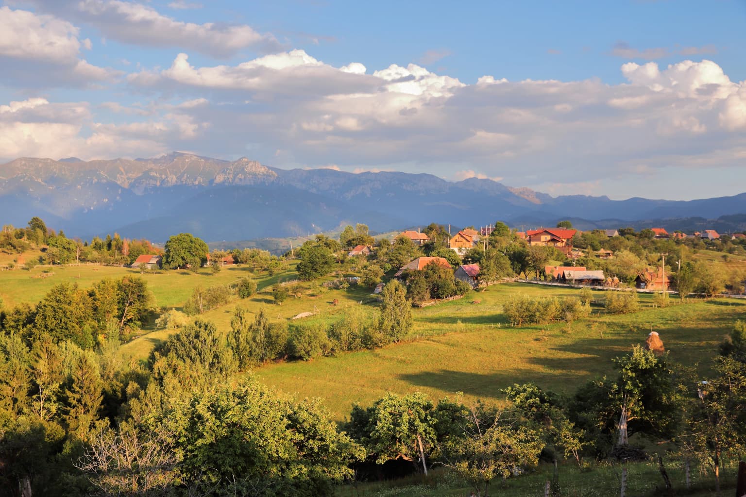 A landscape view of Magura village with Piatra Craiului mountains visible in the background, showing green fields, trees, and scattered houses under a partly cloudy sky
