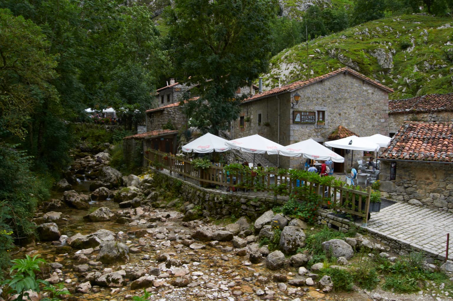 River with rocky bed, stone buildings, white umbrellas, green hills, and trees.