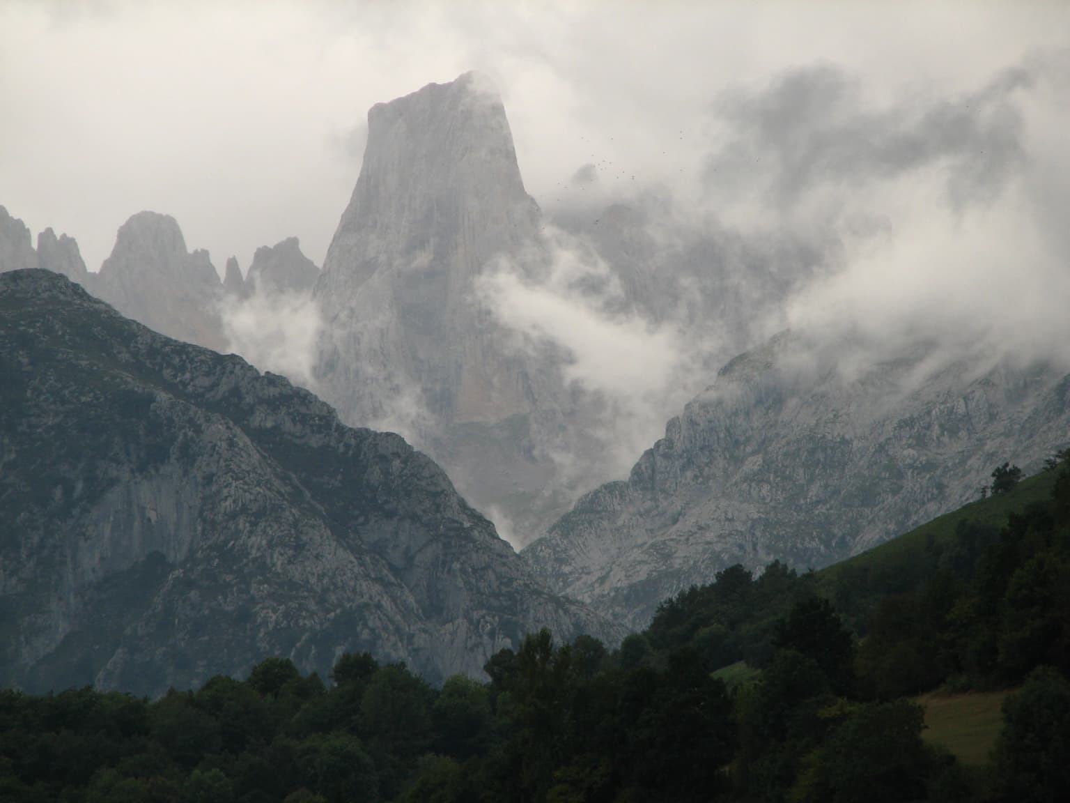 Mountainous landscape featuring a prominent rocky peak partially obscured by clouds, with forested areas in the foreground and rugged rock formations in the background