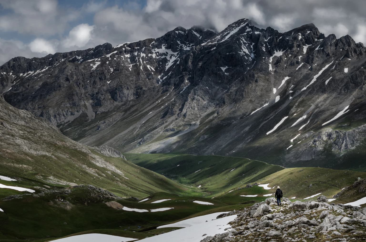 A person standing on rocky terrain with patches of snow, green valleys, and mountain peaks in the background