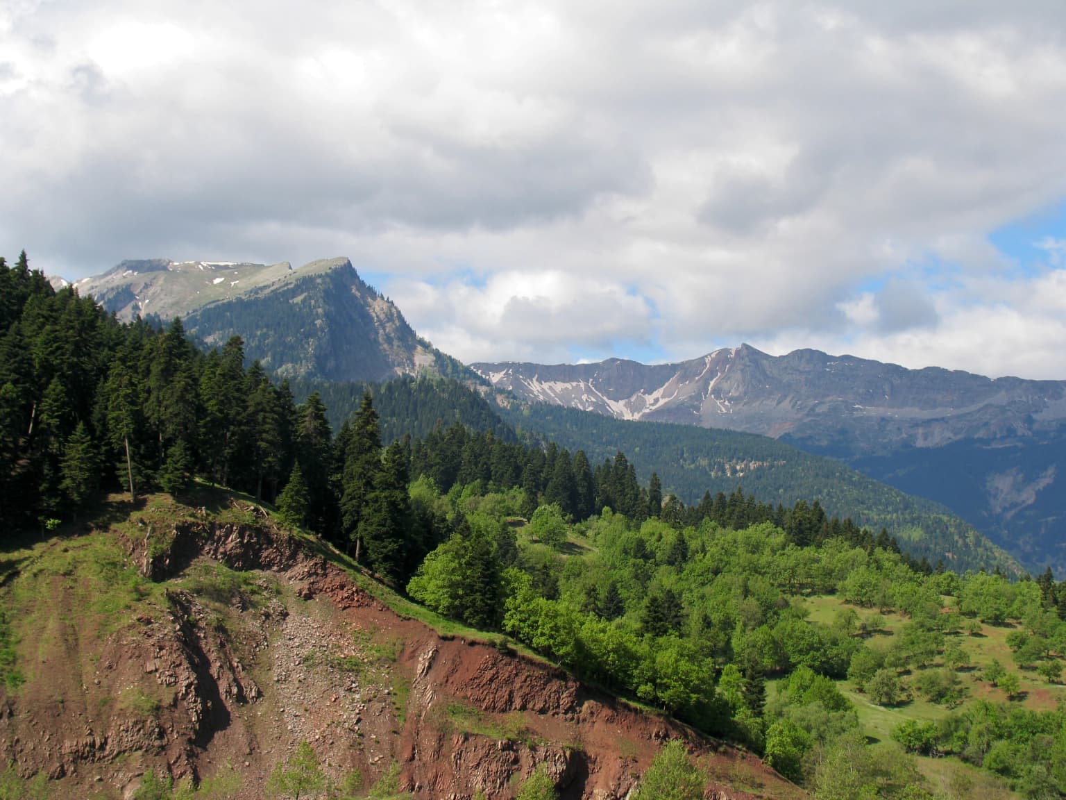 Panoramic view of the Pindos mountain range with dense forests, rocky hills, and distant peaks under a partly cloudy sky.