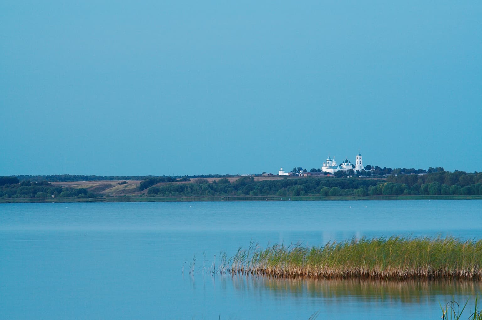 Calm blue lake with tall reeds in foreground and white monastery on distant hill under clear blue sky