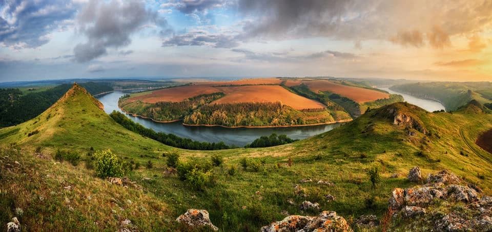 Panoramic landscape of green rolling hills, a winding river, and a valley under a partly cloudy sky