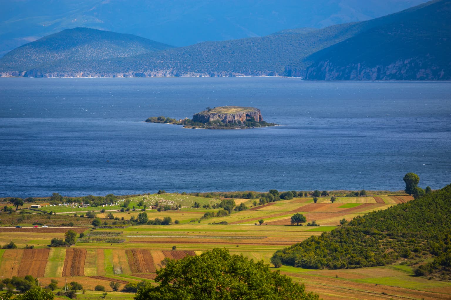 Wide view of Lake Prespa with Mali Grad Island in the center, surrounded by water, with agricultural fields and hills in the foreground and mountains in the background
