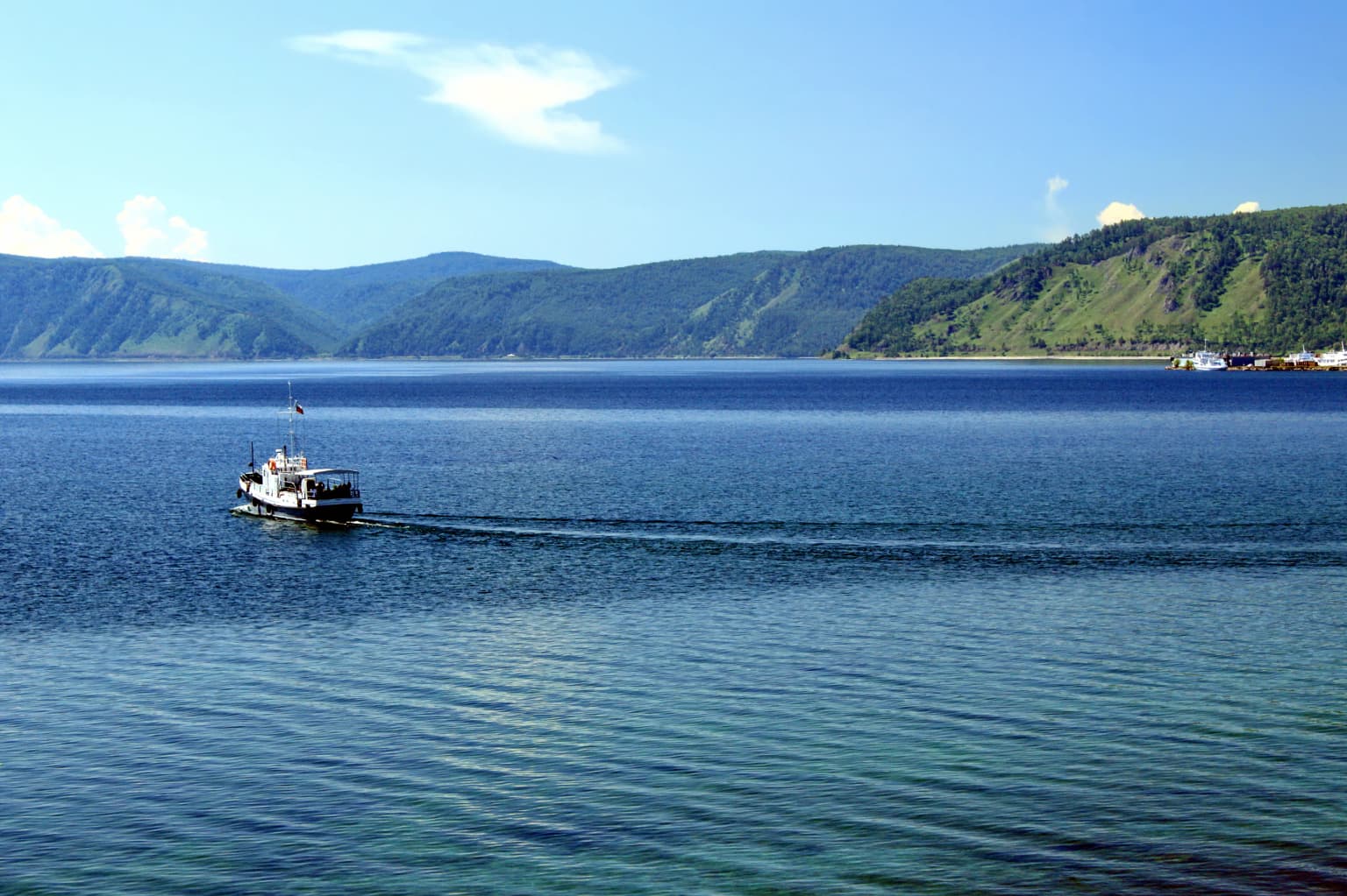 A boat traveling on the calm waters of Lake Baikal with green hills and a clear blue sky in the background