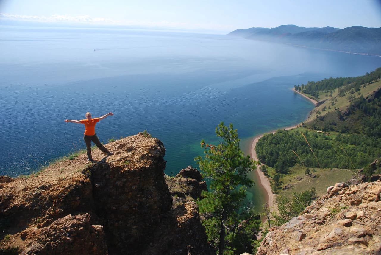 Person in orange shirt standing on rocky cliff with arms outstretched overlooking blue lake and coastal road