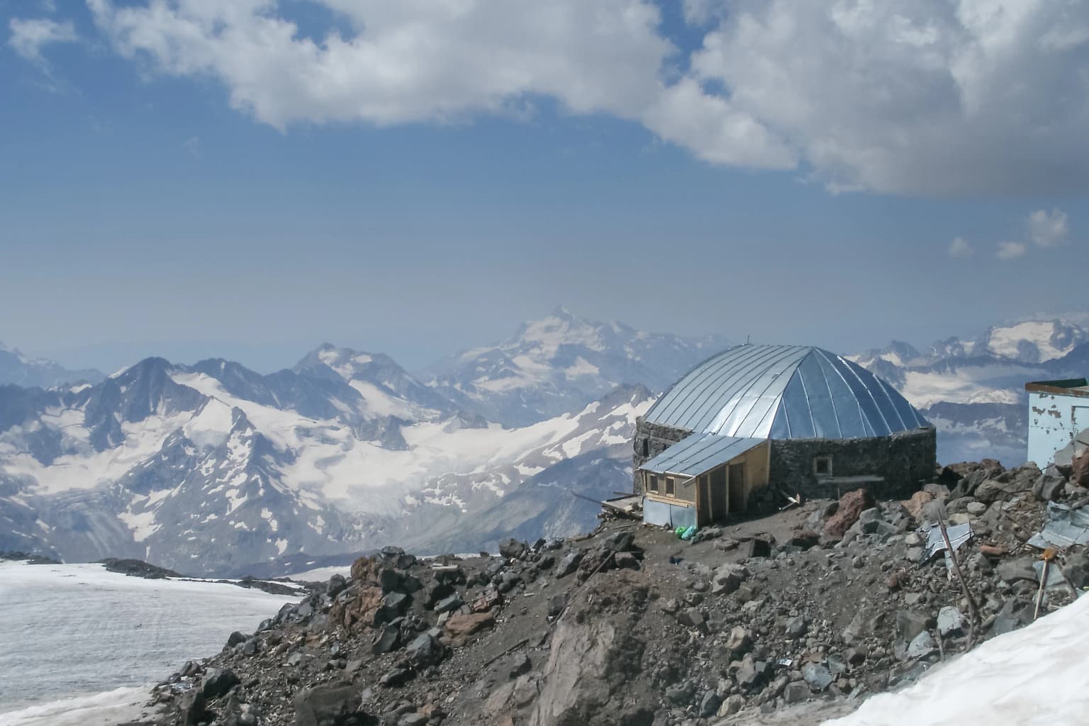 Metal-roofed mountain shelter on rocky terrain with snow-covered peaks in background
