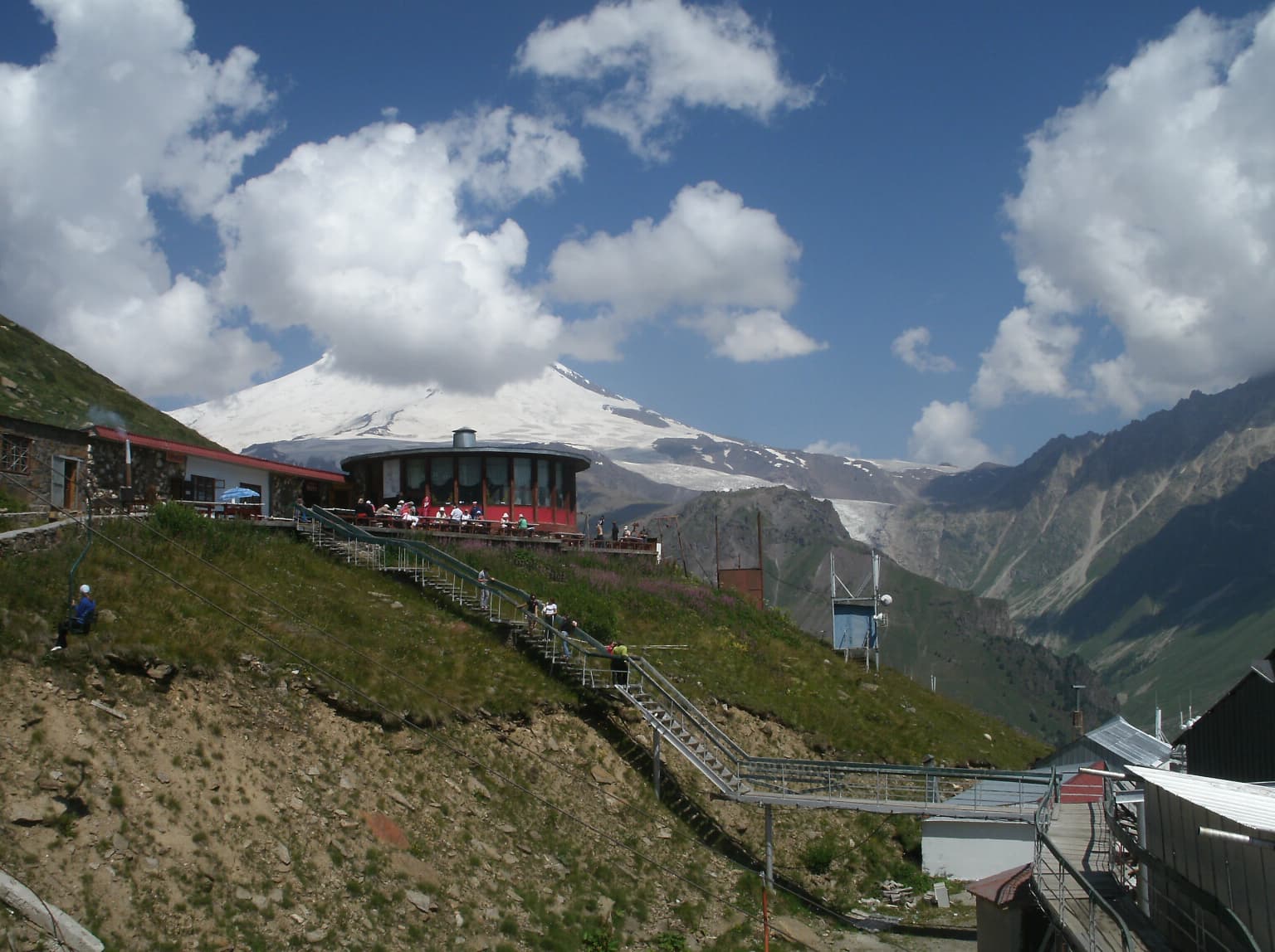Circular building on grassy hillside with people walking on stairs, Mount Elbrus snow-capped peak visible in background
