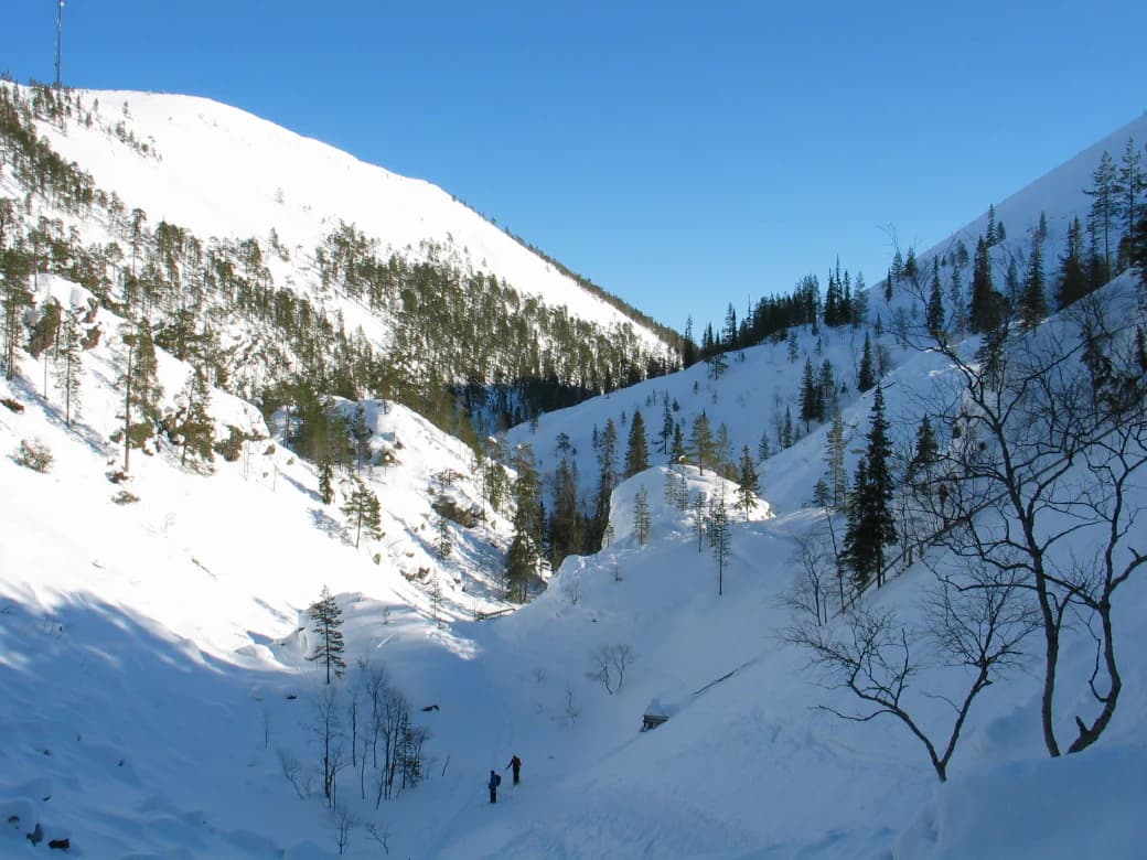 Two people walking through a snow-covered valley between mountain slopes with scattered pine trees under clear blue sky