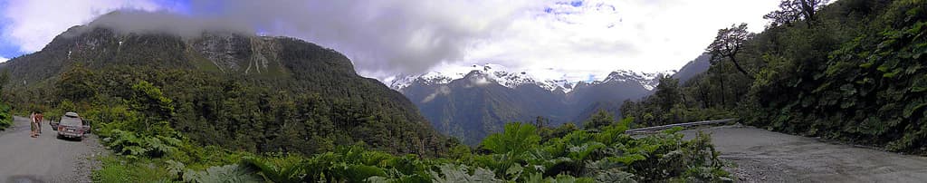 Vista panorámica de una carretera serpenteante a través de densa vegetación verde con montañas nevadas a lo lejos, un coche en la carretera y una persona caminando