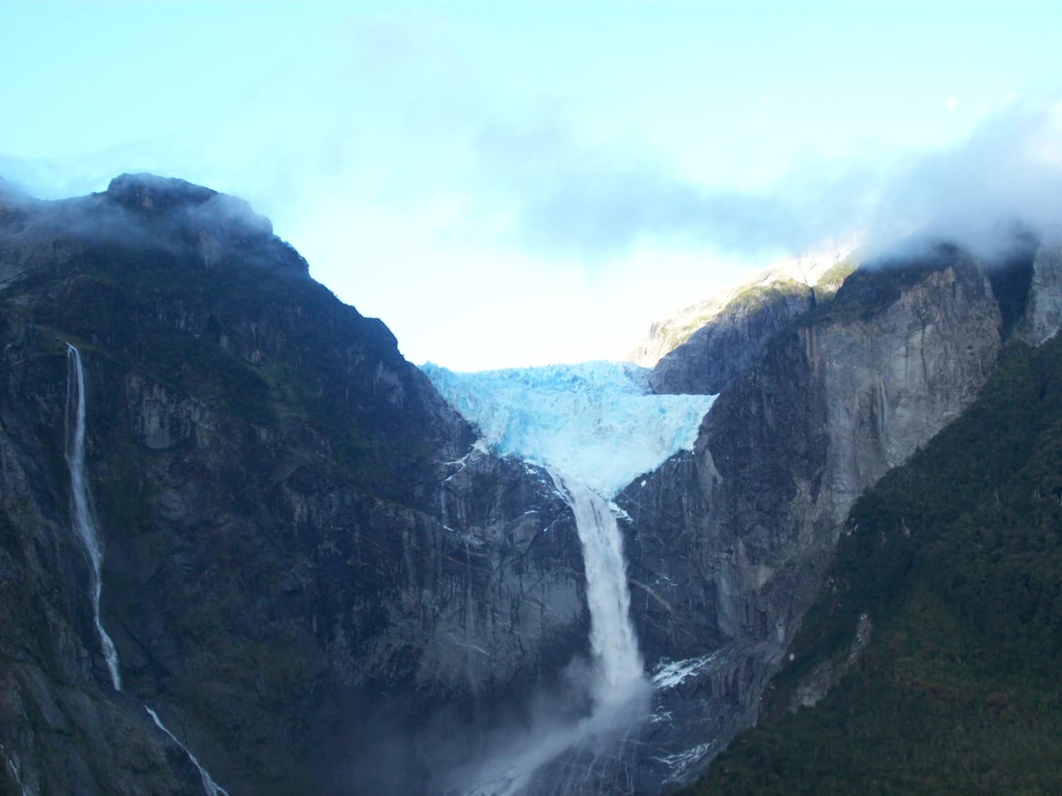 Glaciar colgante en el Parque Nacional Queulat, rodeado de escarpadas montañas rocosas con cascadas cayendo bajo un cielo parcialmente nublado