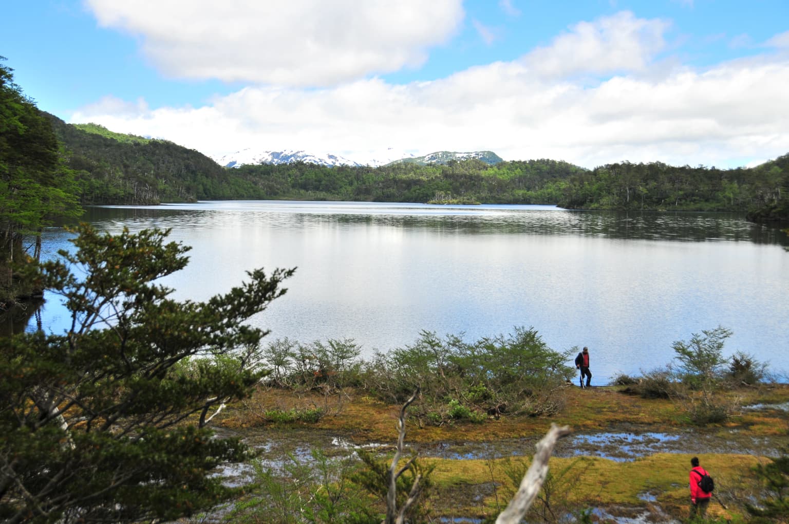 Un lago rodeado de colinas boscosas con montañas al fondo. Dos personas están de pie cerca de la orilla.