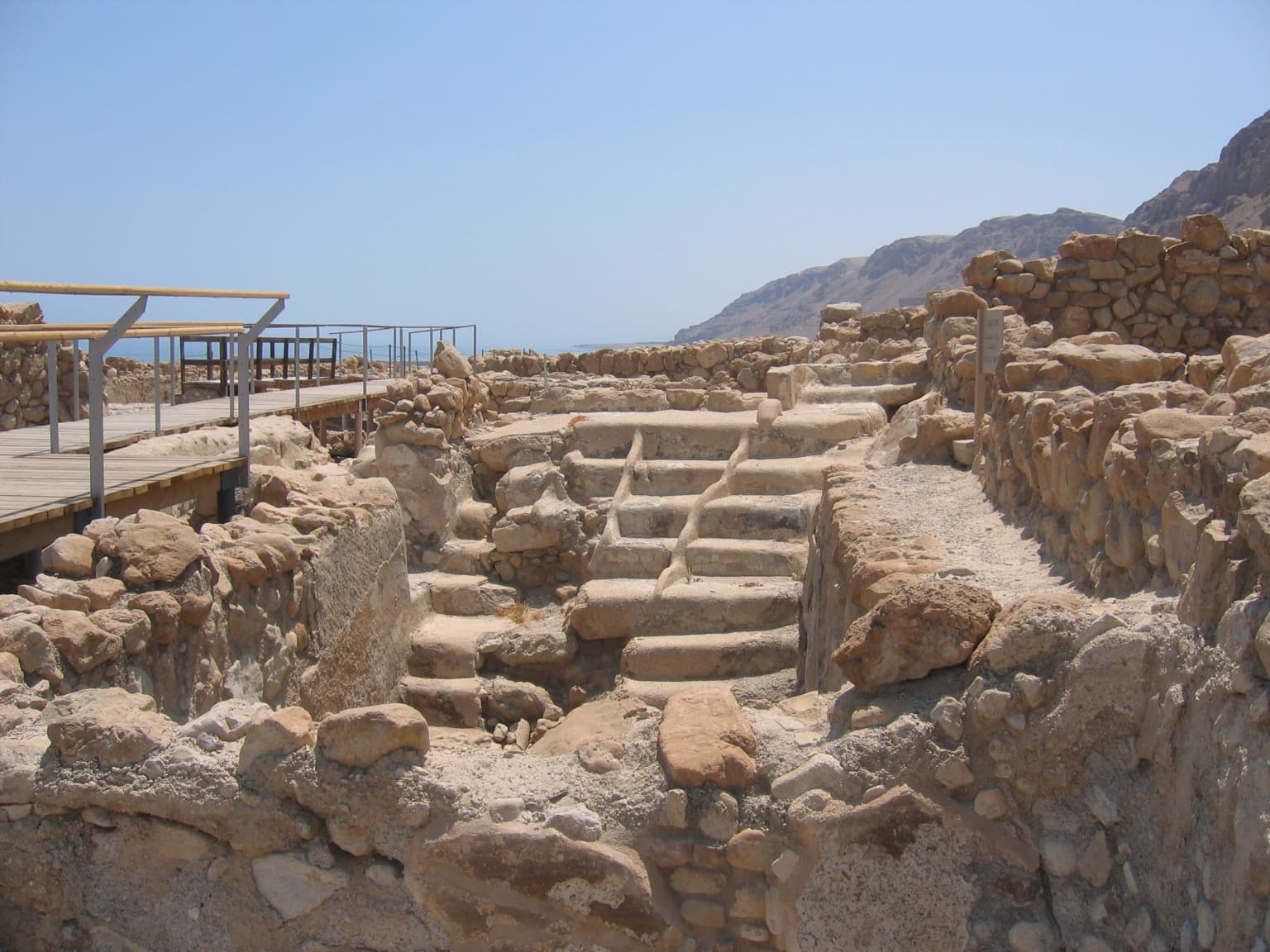 Estructura de baño ritual de piedra con escalones rodeada de antiguos muros de piedra, pasarela de madera con barandillas, montañas y el Mar Muerto visibles al fondo