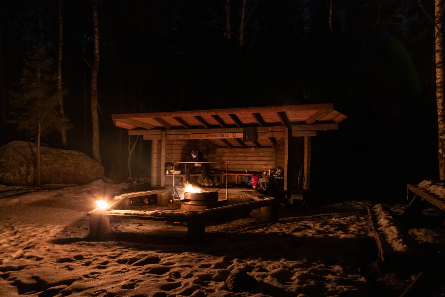 Wooden lean-to shelter with fire pit at night, surrounded by snow-covered ground and trees