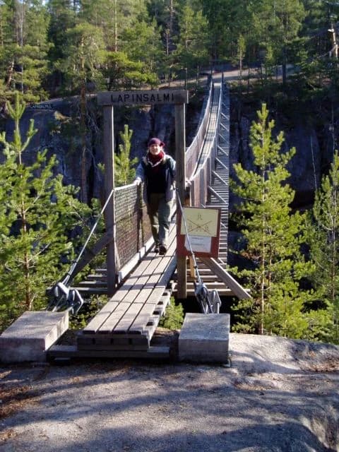 Suspension bridge with wooden planks and metal railings spanning a rocky valley, a person walking on it, surrounded by pine trees.