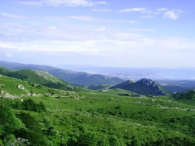 Green mountainous landscape with rolling hills, distant mountains, and a visible sea horizon with islands under a partly cloudy sky