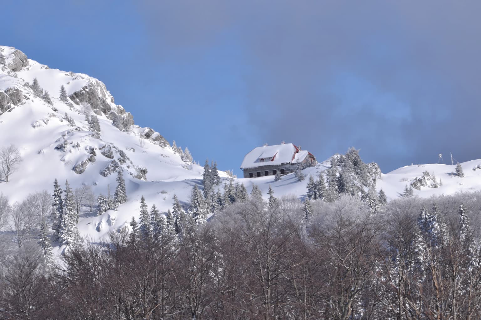 Snow-covered mountain landscape with a building, snow-laden trees, and rocky peaks under a partly cloudy sky