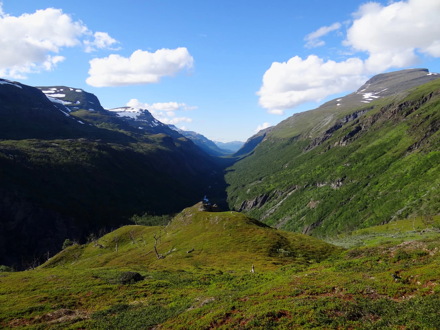A wide valley with steep green slopes and snow-capped mountains in the distance under a blue sky with scattered clouds.