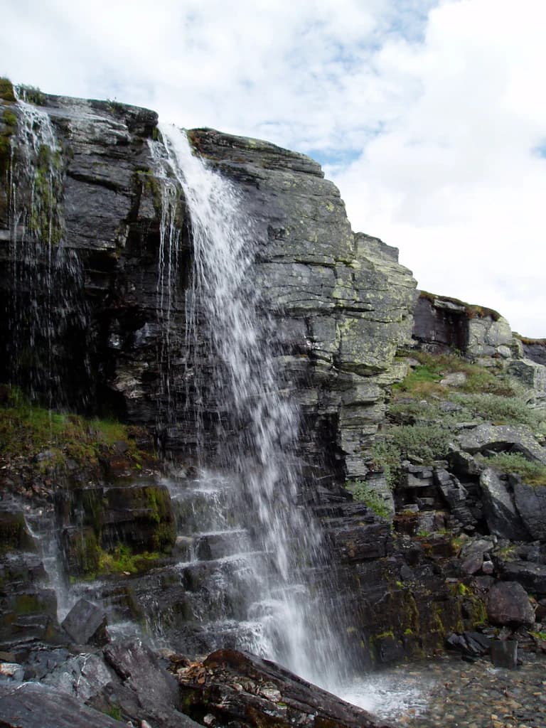 Waterfall flowing down rocky cliffs with moss-covered rocks and scattered boulders under a partly cloudy sky