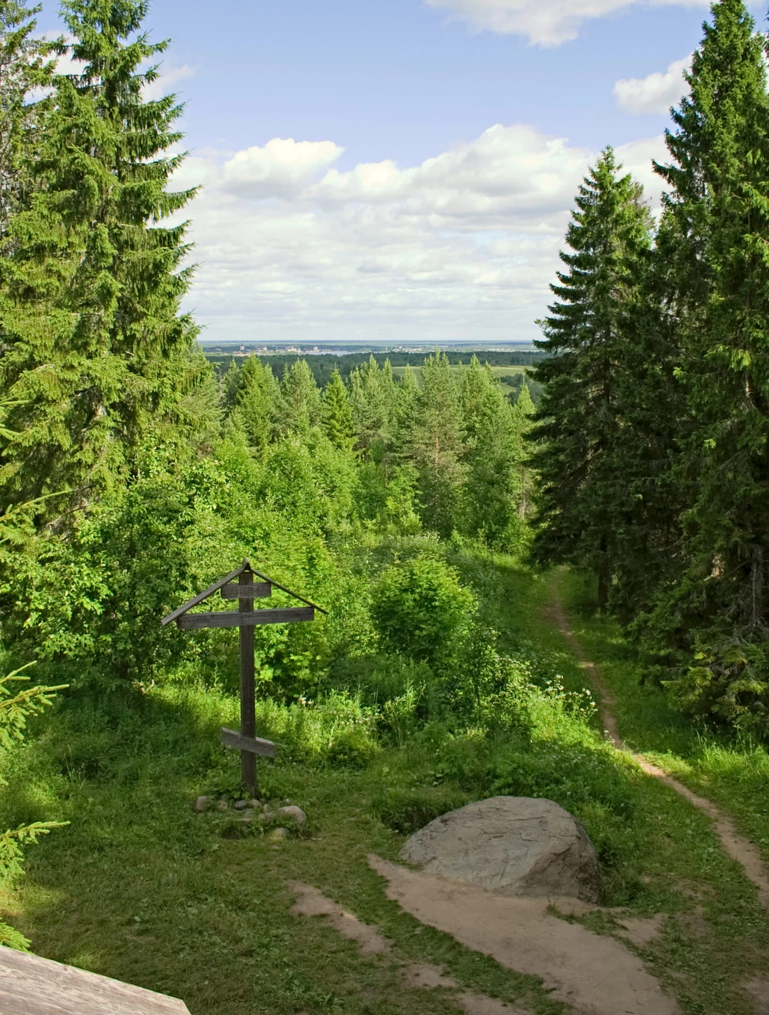 Wooden cross on a grassy hill, surrounded by tall evergreen trees, with a dirt path and distant view of the town of Kirillov.