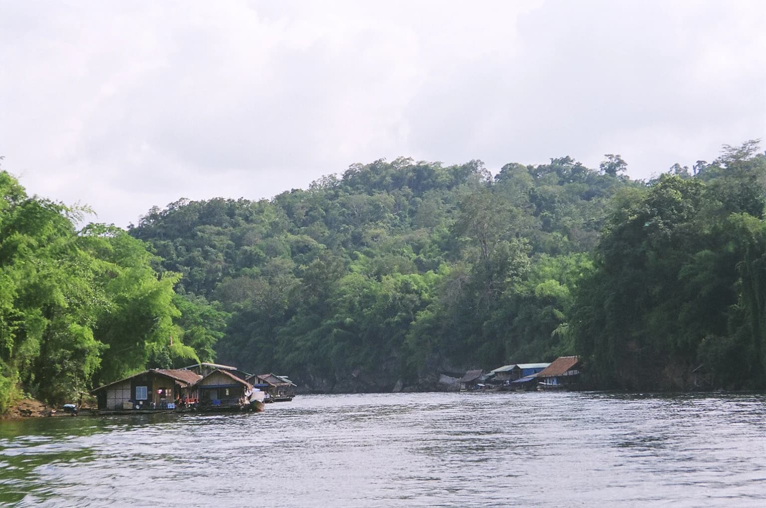 A wide river with calm water bordered by dense green forests and small riverfront structures under a partly cloudy sky.