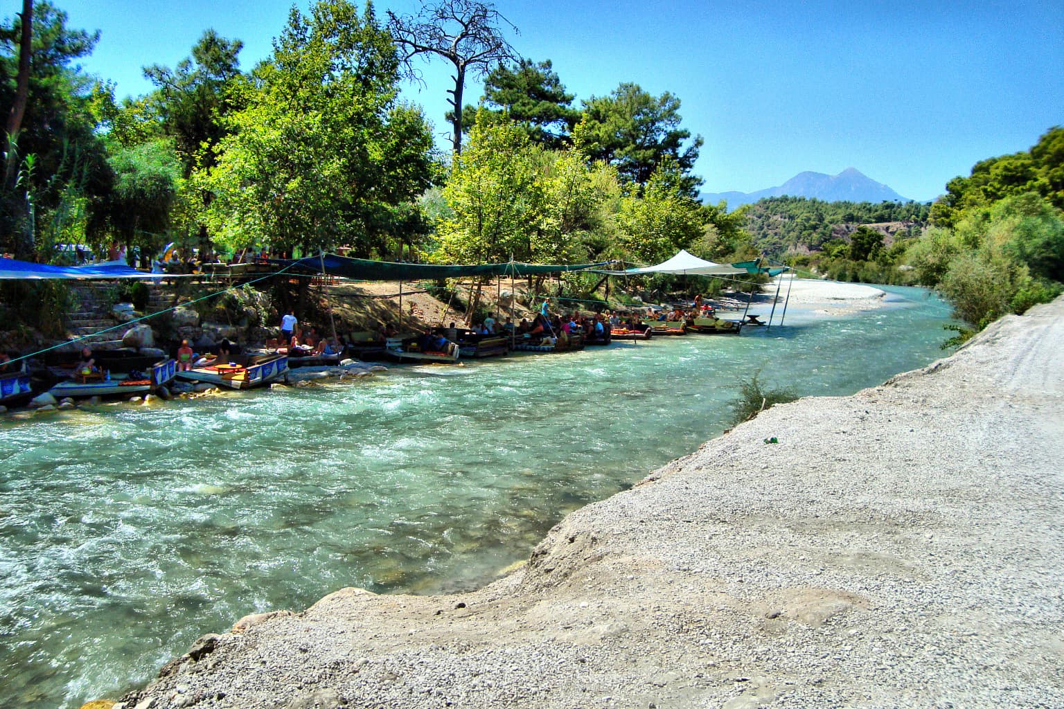 Wide river flowing through canyon with rafts, trees, and bridge under clear blue sky
