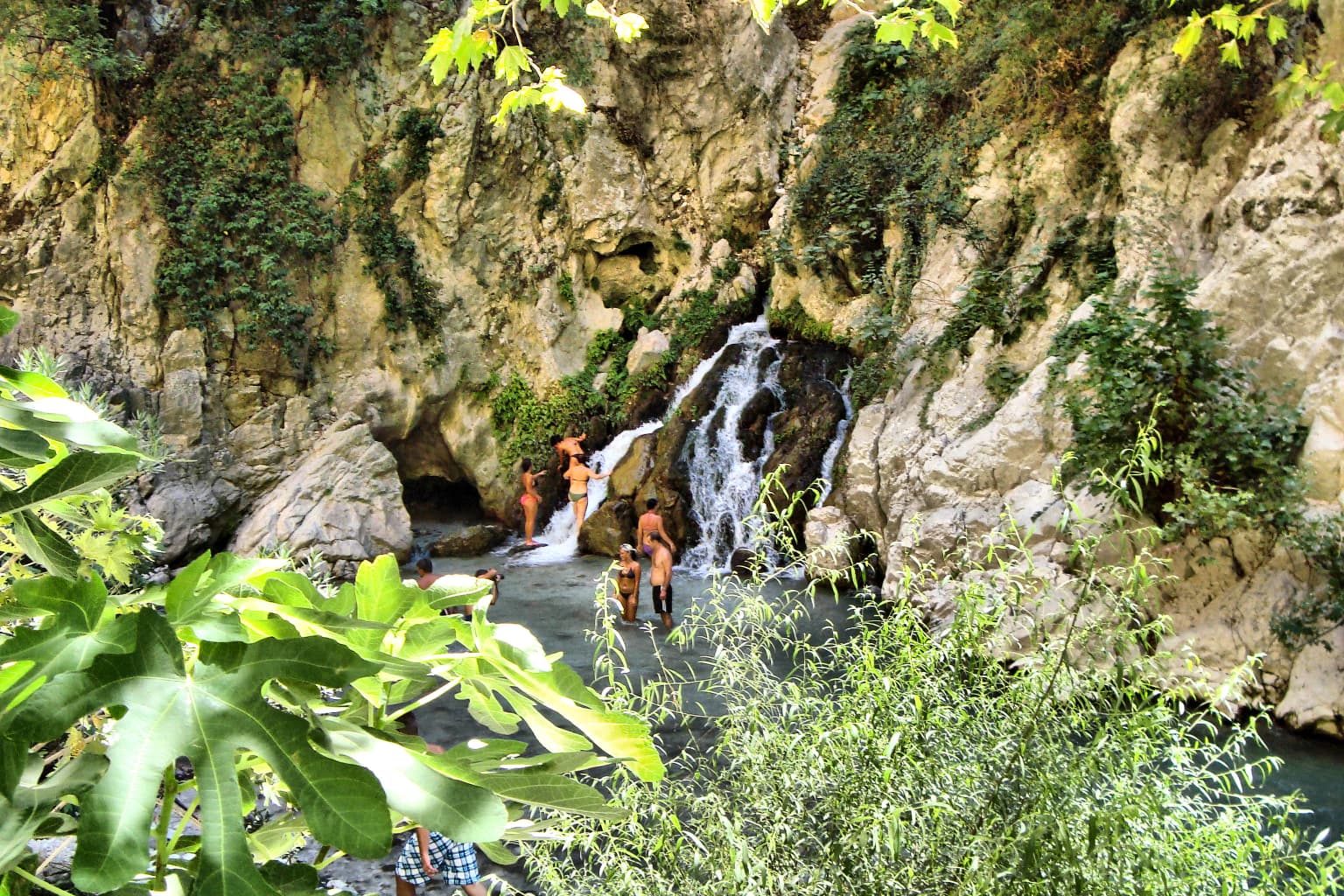 Waterfall cascading over rocky cliffs with visitors swimming in a pool below, surrounded by green vegetation.