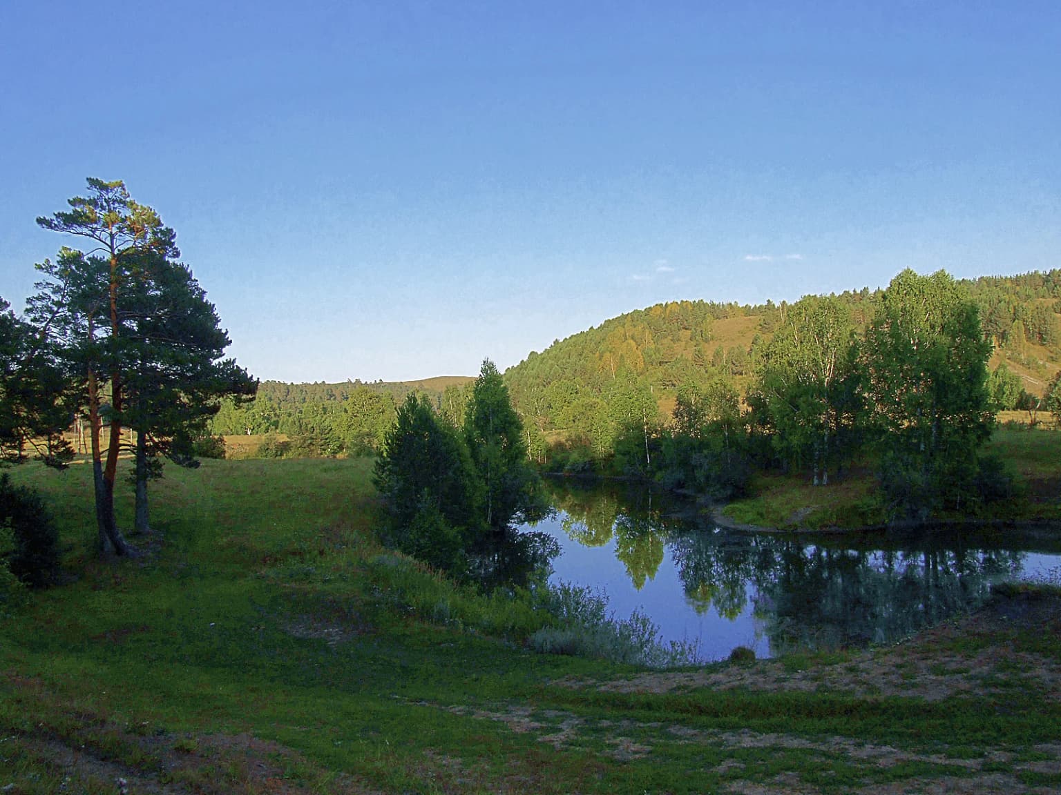 A serene lake reflecting green trees and hills in a forested landscape with a clear blue sky