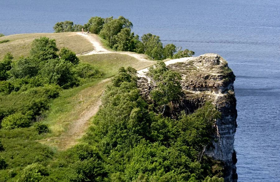 Cliff with a dirt path winding through green vegetation, overlooking a body of water