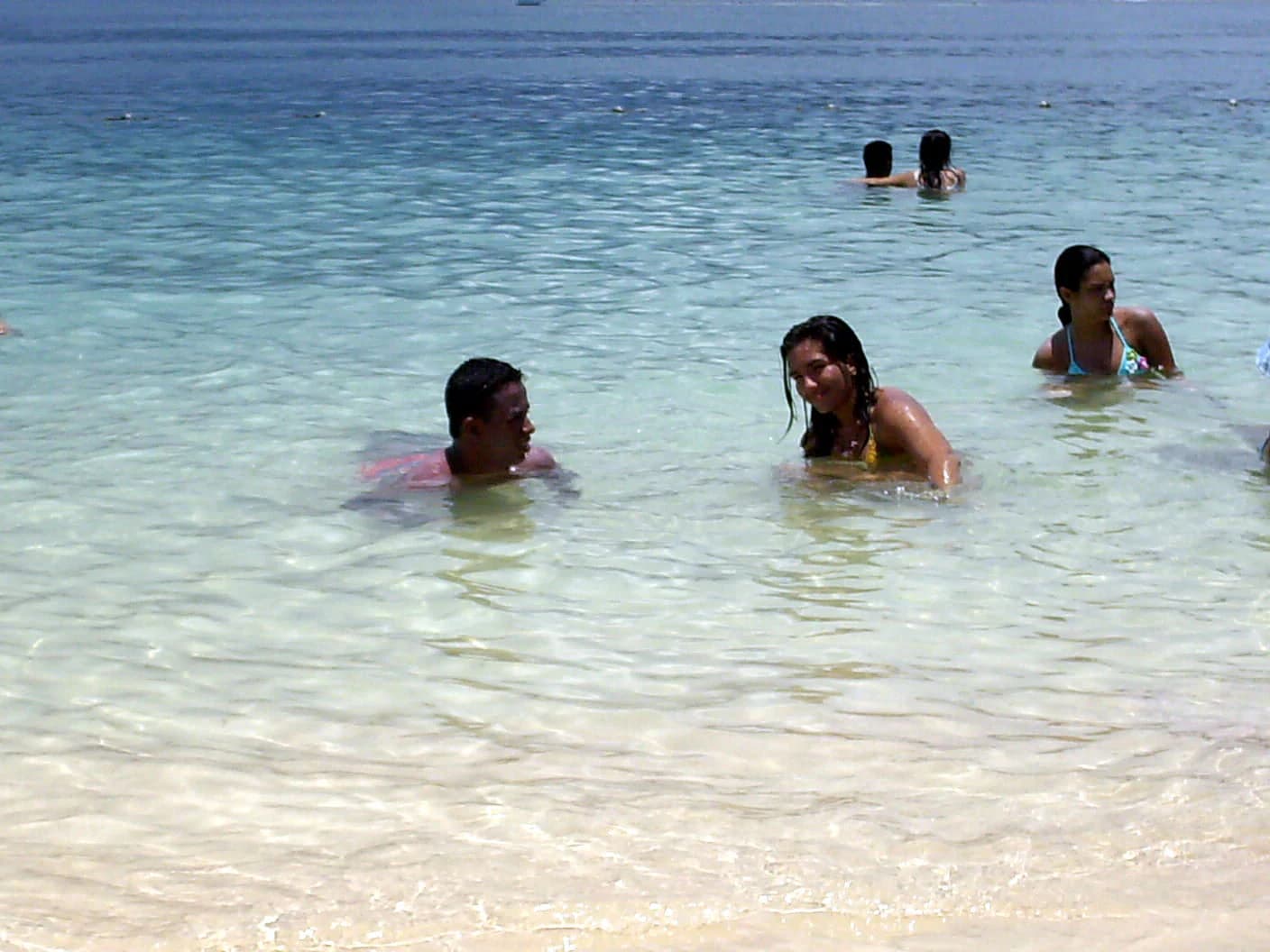 Personas nadando y vadeando en aguas turquesas poco profundas en una playa de arena