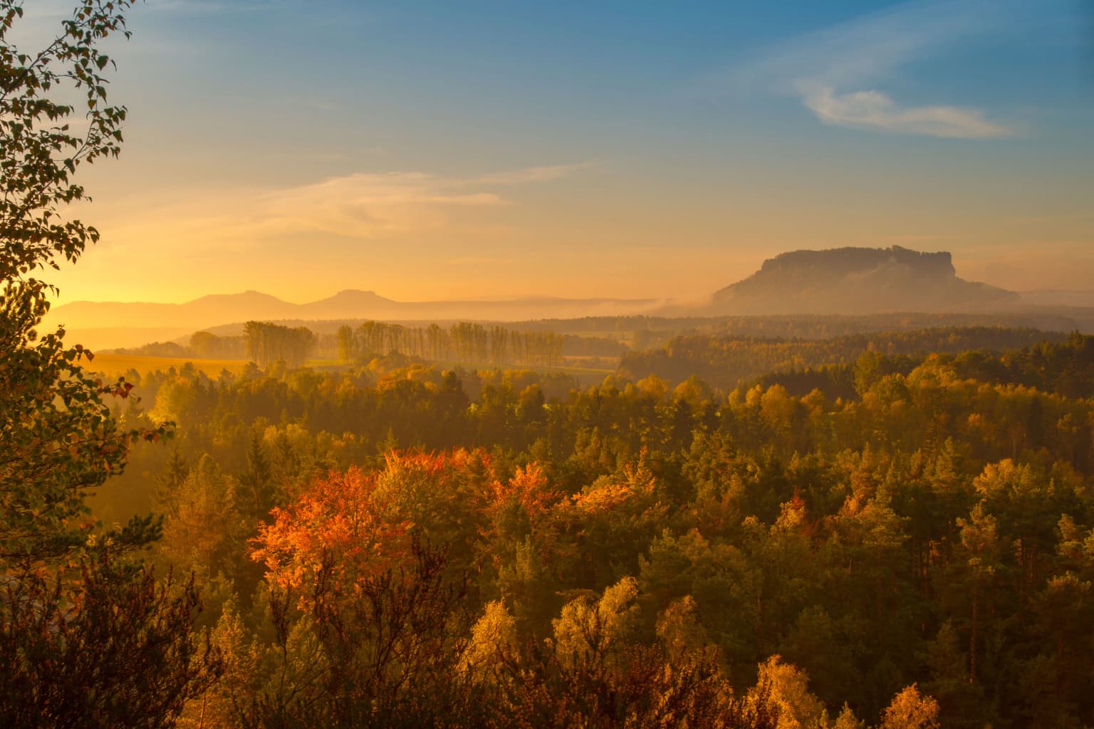 Landscape view of Lilienstein rock formation at sunrise with forested hills and distant mountains under a clear sky