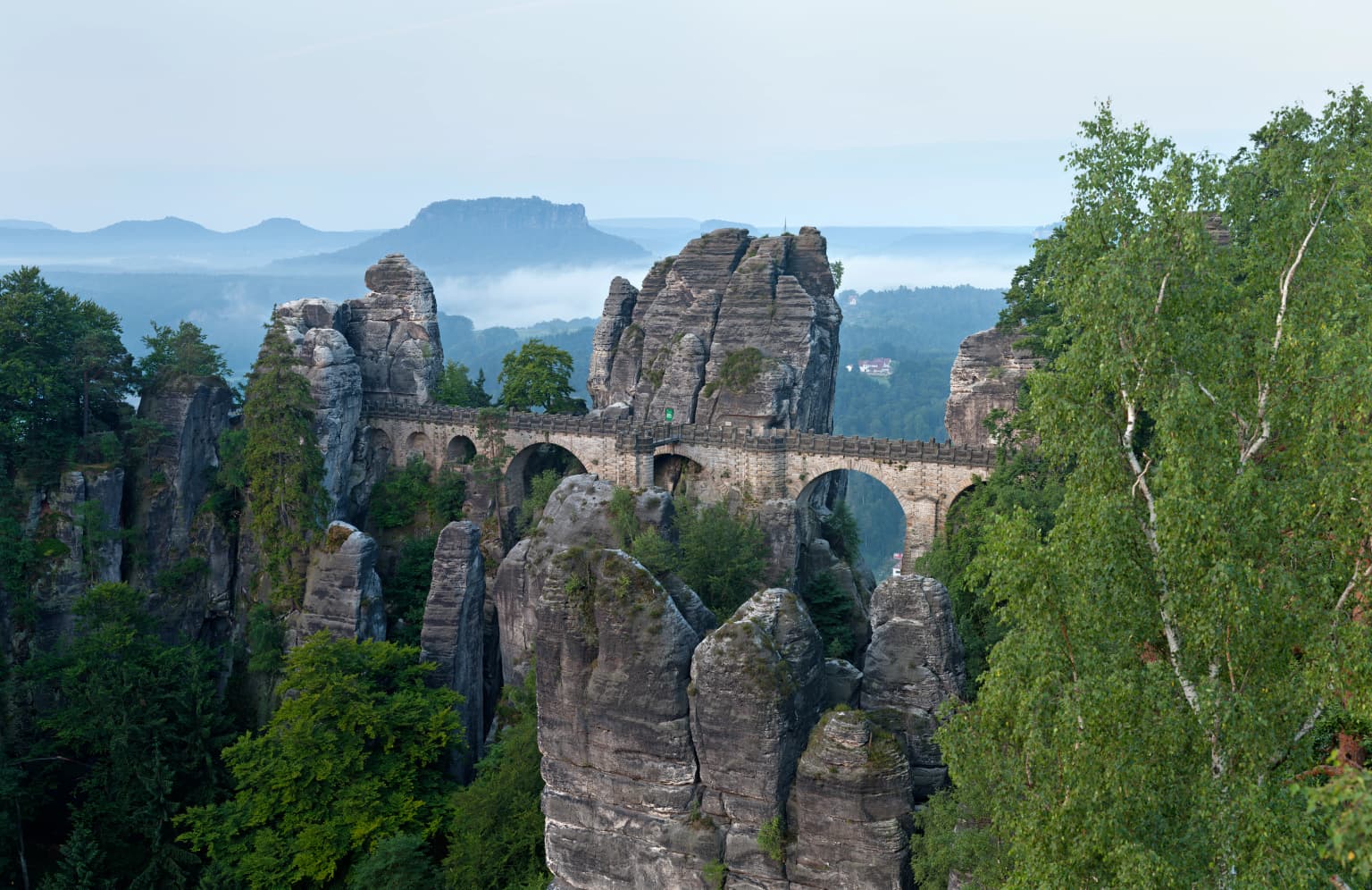Stone arch bridge connecting two sandstone cliffs with forested mountains and misty horizon in background