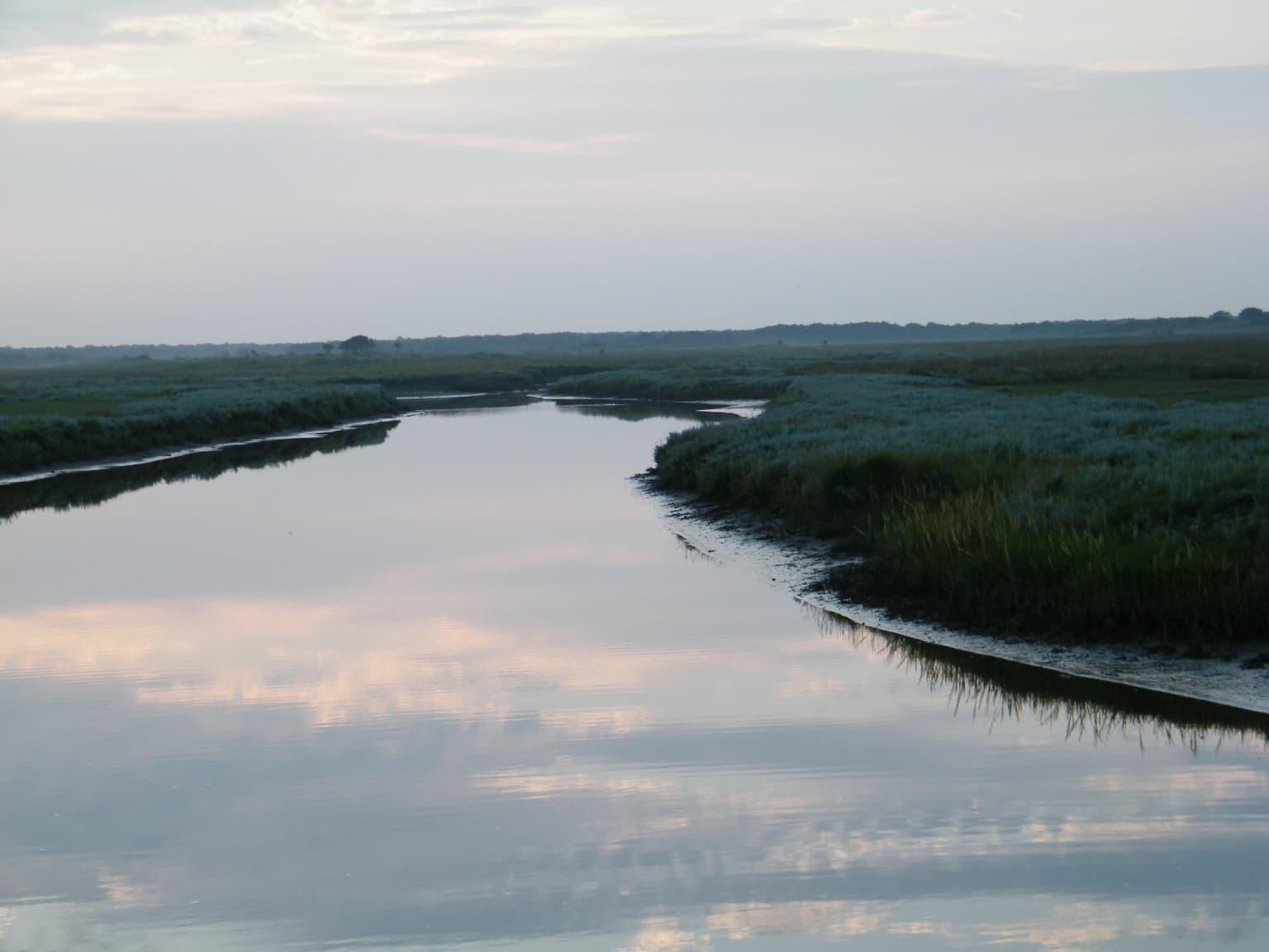 A calm waterway with reflections of the sky, bordered by grassy wetlands
