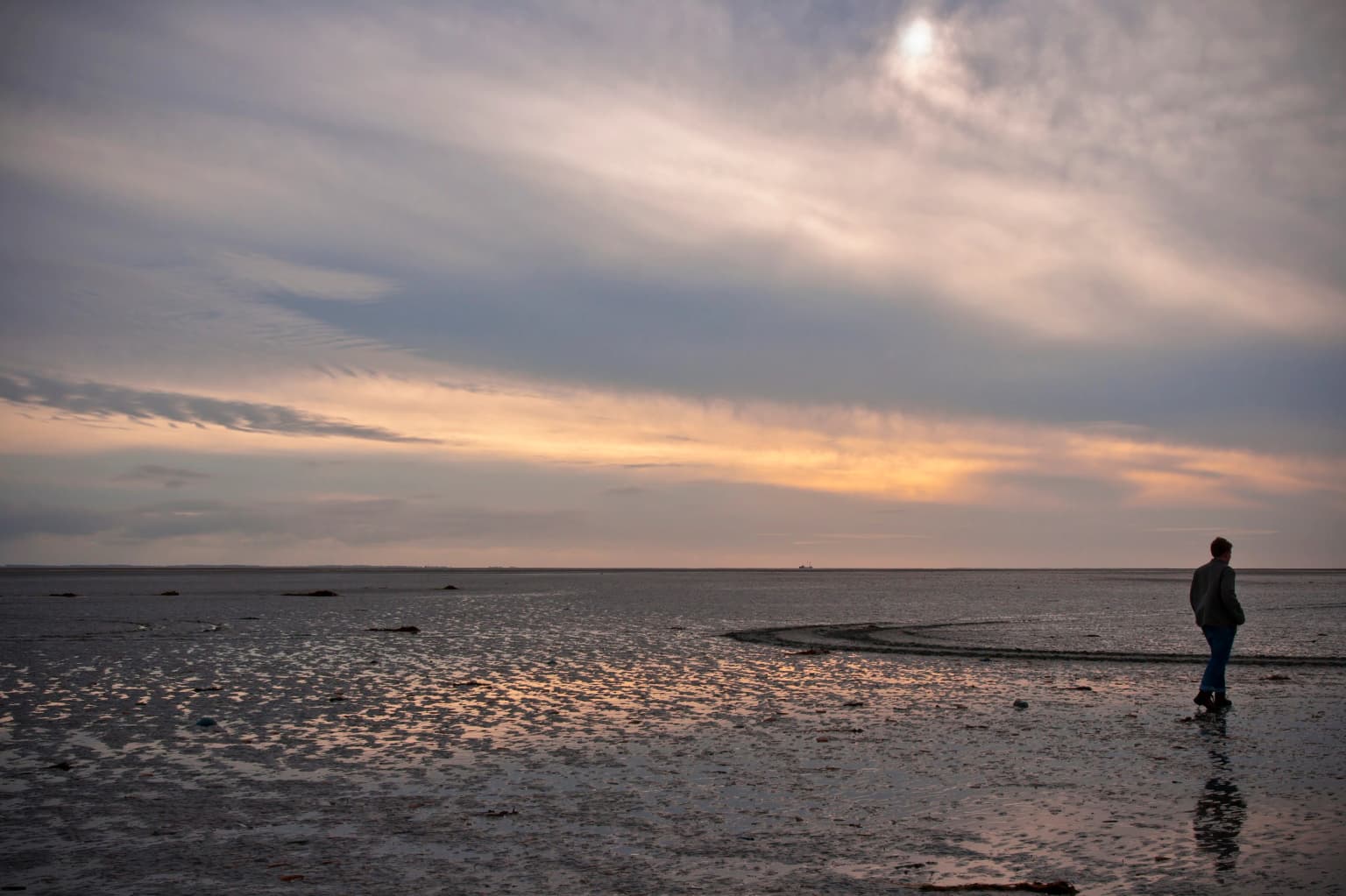 A person standing on wet sandy flats under a partly cloudy sky with sunset colors