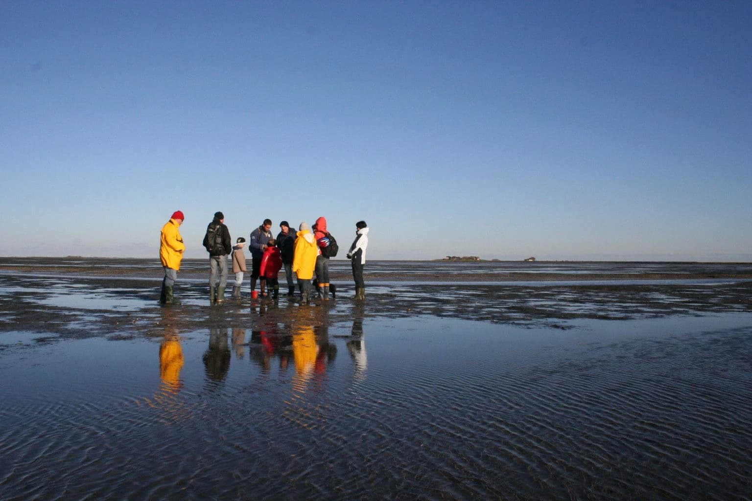 Group of people standing on a reflective mudflat under clear blue sky with visible reflections in wet sand.