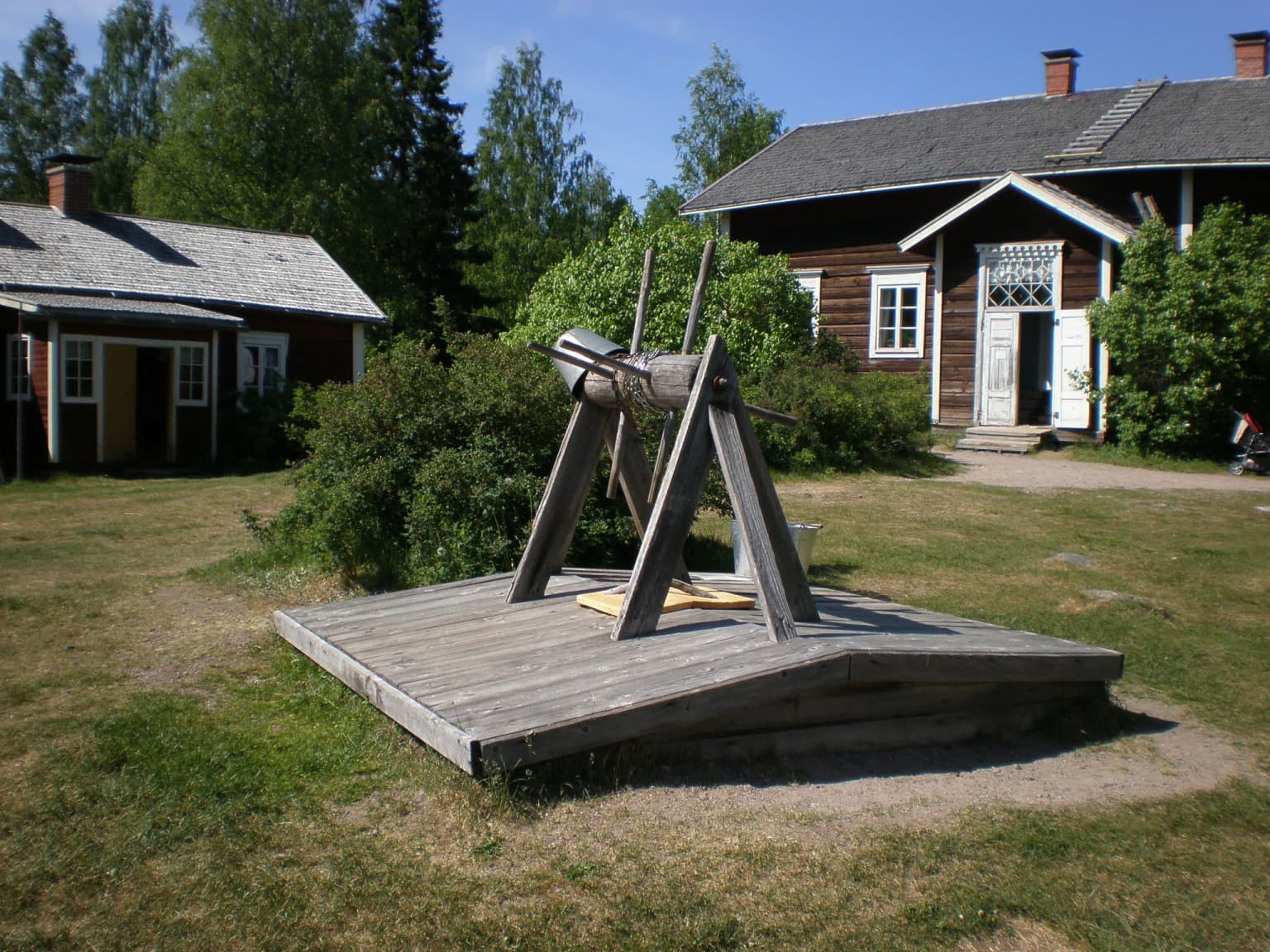 Wooden farm implement structure on a platform with two traditional houses and greenery in the background