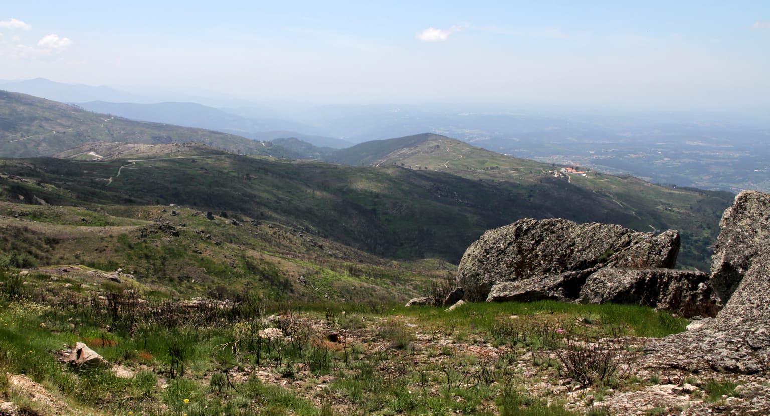 Mountain landscape with rocky formations, rolling green valleys, and distant hills under a clear sky