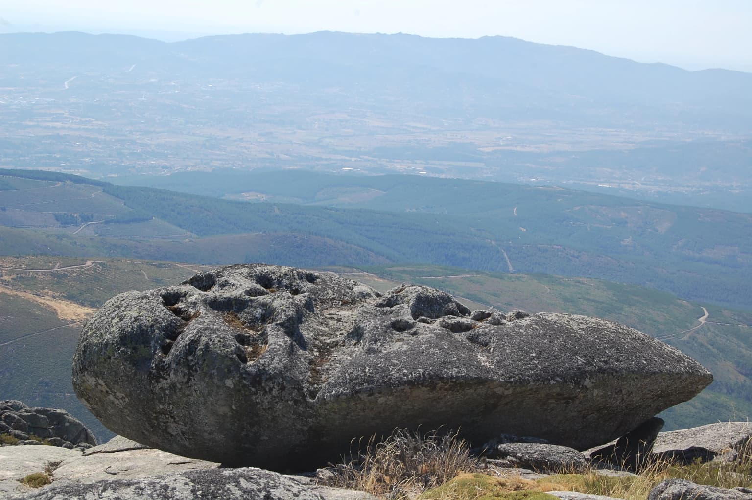 A large, uniquely shaped rock formation with holes sits on a mountain peak with panoramic views of valleys and distant mountains.