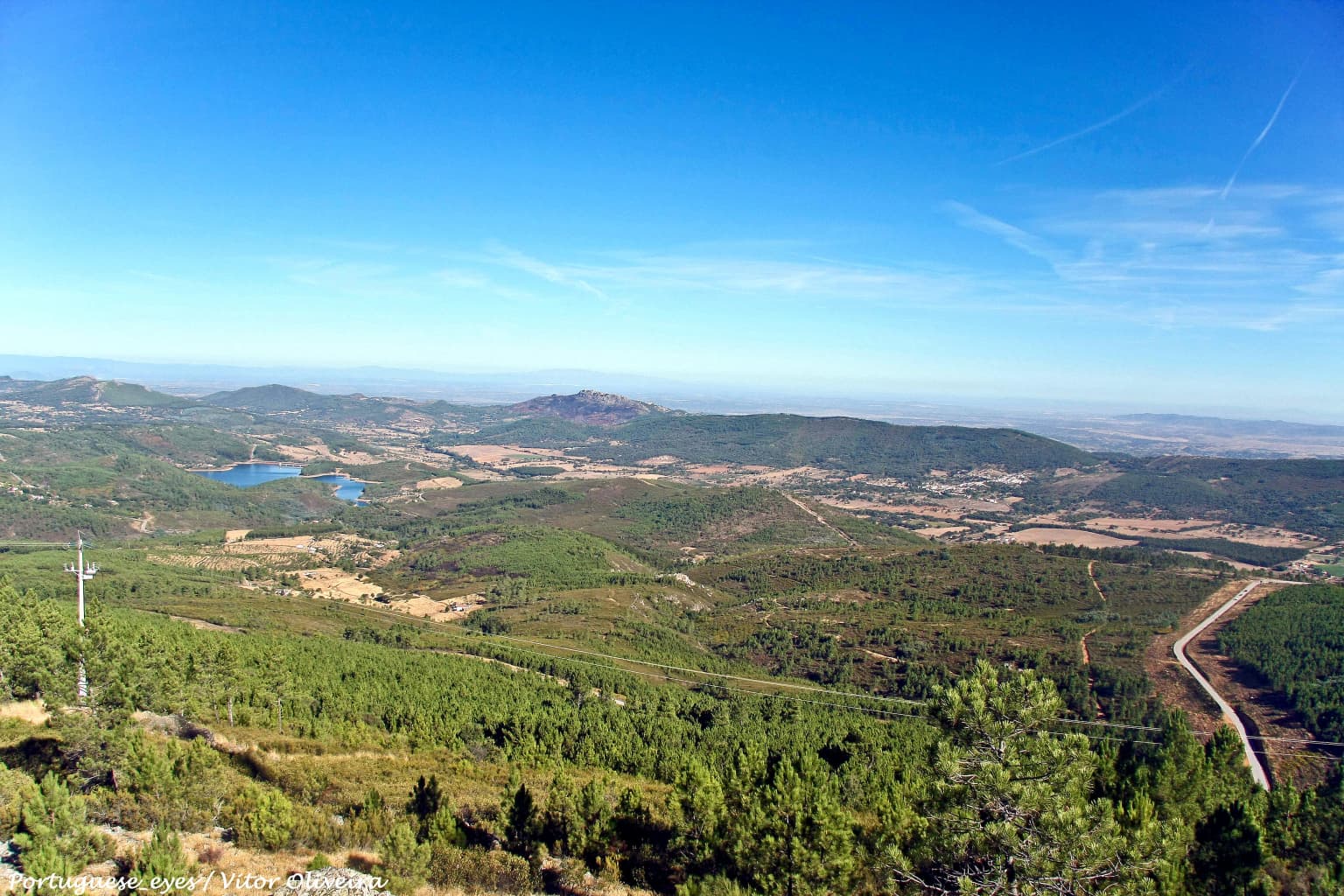 Wide landscape view from a high vantage point showing rolling hills, forests, a small lake, and a winding road under a clear blue sky