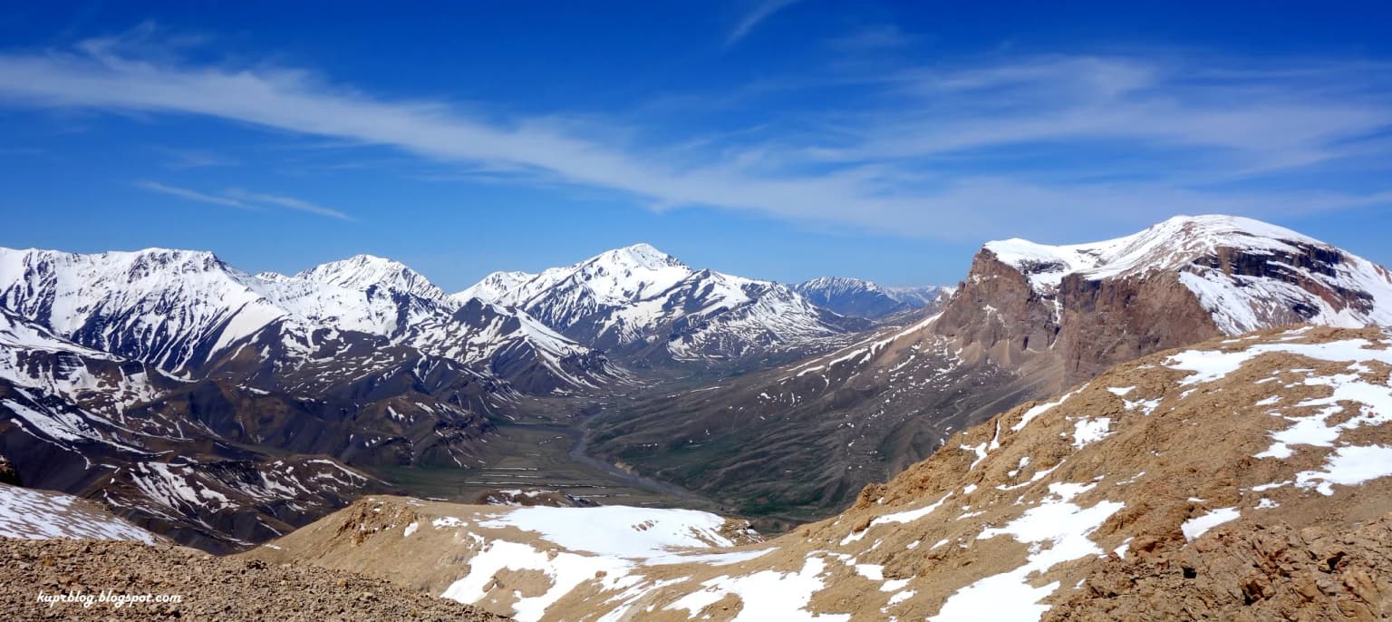 Wide-angle view of a mountain valley with snow-covered peaks, rocky terrain, and clear blue sky