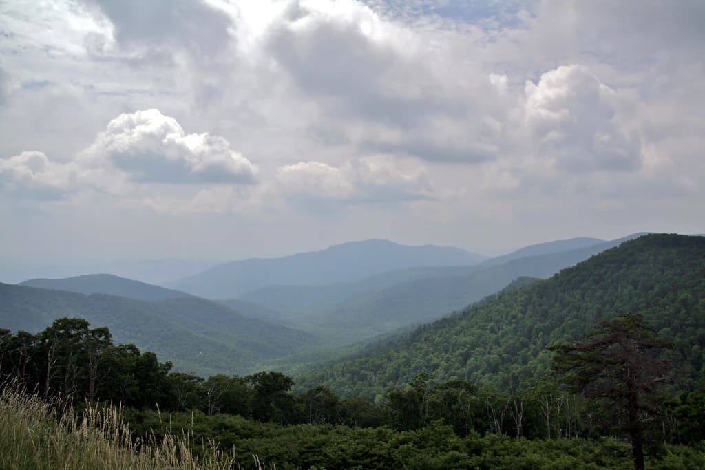 Wide landscape view of green mountain ridges, forested valleys, and distant hills under a partly cloudy sky