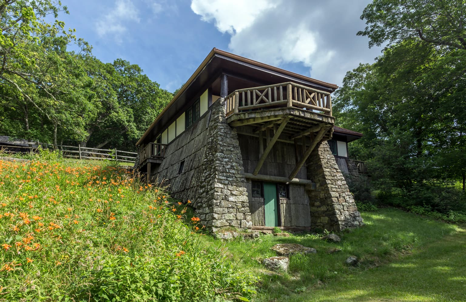 Edificio de un albergue de piedra y madera con balcón de madera, rodeado de césped verde y flores silvestres anaranjadas, con árboles al fondo
