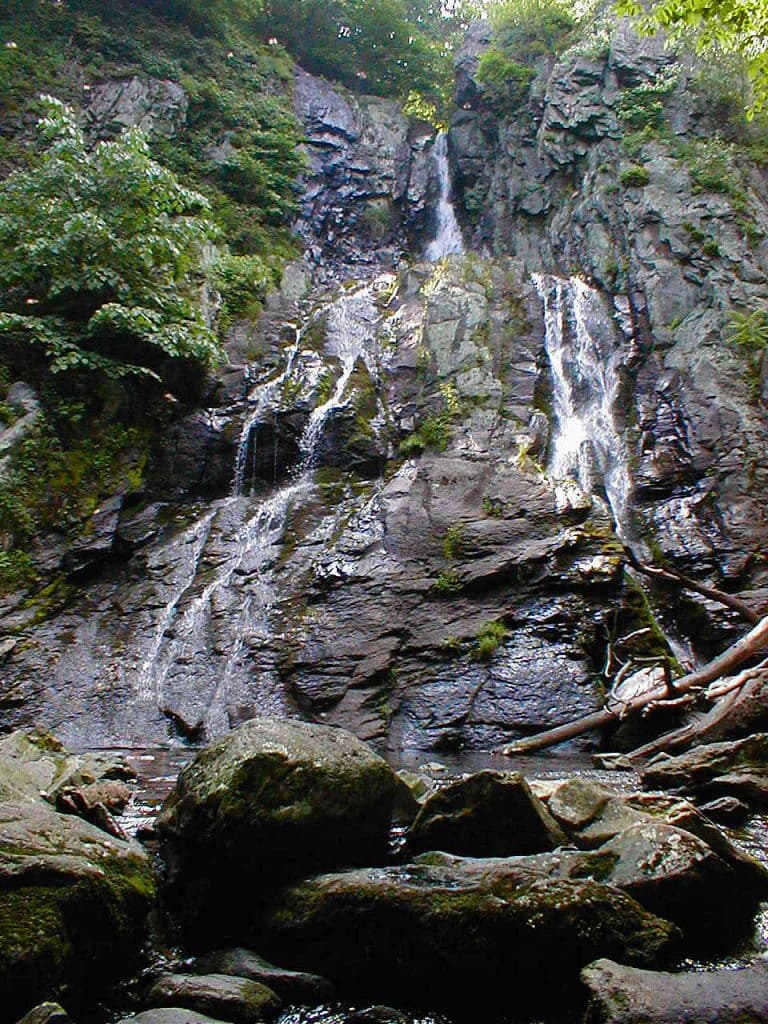 Waterfall with multiple streams flowing over moss-covered rock face, surrounded by green vegetation and large boulders