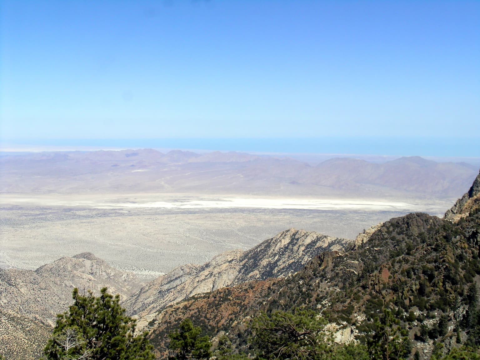 Mountain landscape with pine trees and rocky slopes in foreground, extending to a valley and distant mountain ranges under clear blue sky