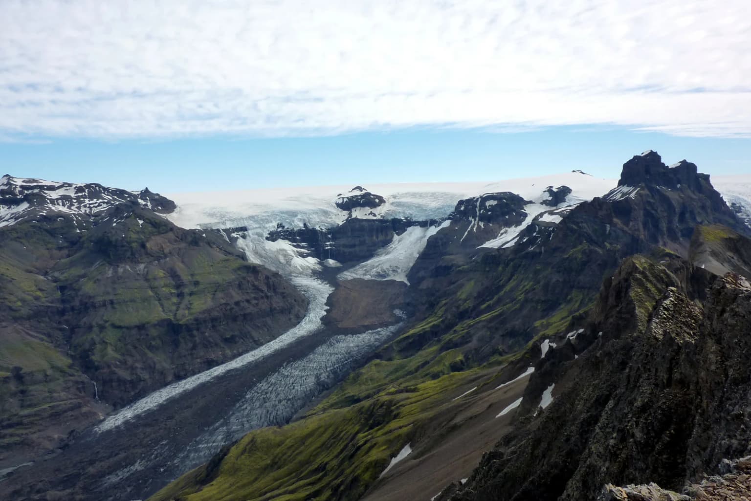 Panoramic view of a glacier flowing through a mountain valley with rocky peaks, grassy slopes, and snow patches under partly cloudy sky