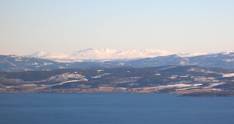 Wide landscape view showing a large body of water with forested hills in the midground and snow-capped mountains in the background under a clear sky