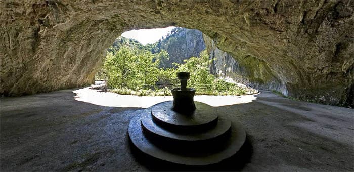 Stone cave entrance with tiered stone fountain, opening to sunlit valley with green trees and rocky cliffs