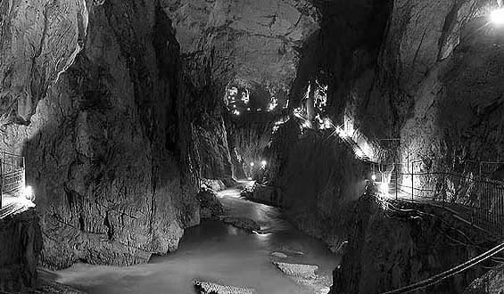 Black and white photograph of a cave interior showing a flowing river with rocky walls and artificial lighting along a walkway
