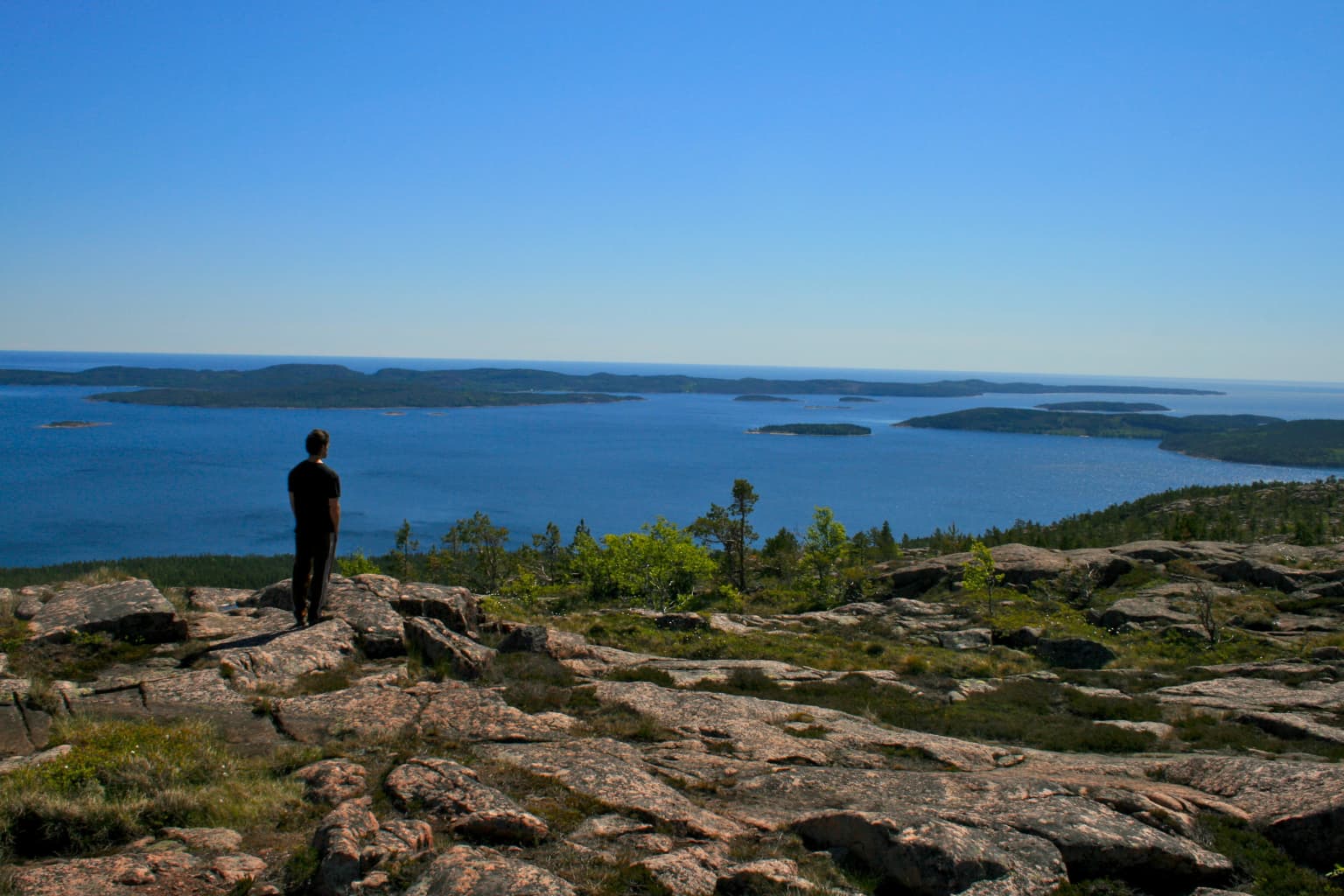 A person standing on a rocky hilltop overlooking a large body of water with distant islands under a clear blue sky.