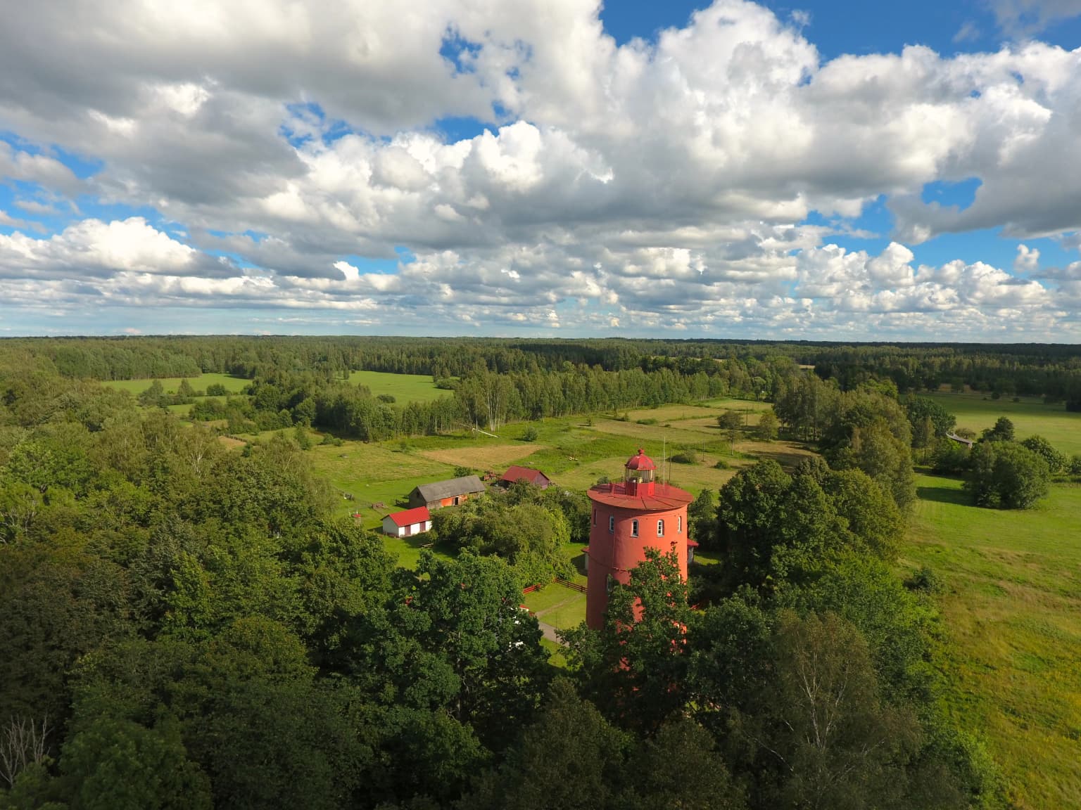 Aerial view of a red lighthouse surrounded by dense green forests with open meadows and small buildings under a partly cloudy sky